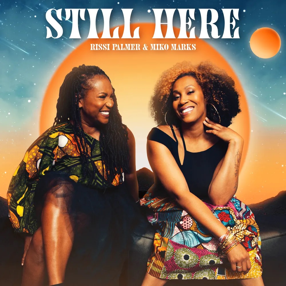 A promotional image of Pissi Palmer and Miko Marks from their "Still Here" single. The two women are sitting together against a stylized orange and blue background.