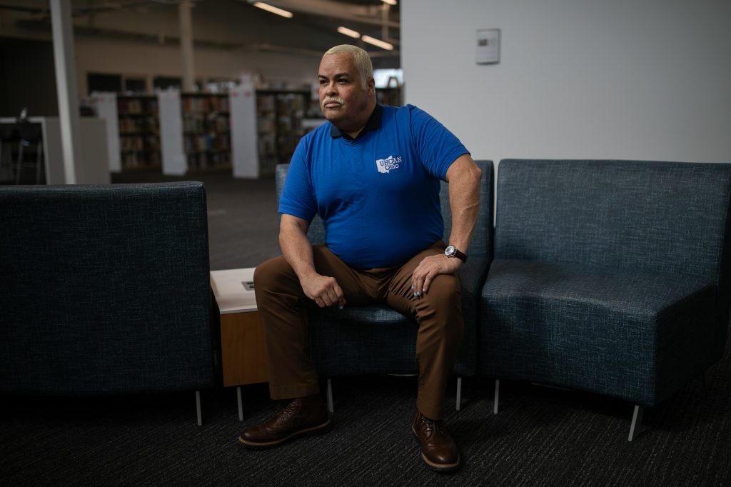 Camacho poses for a portrait sitting down in a library. They have one hand on a knee and their other arm braces on the other knee.