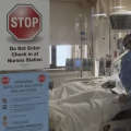 A nurse stands at the bed of a patient in a hospital. Signs on the window warn about precautions against infection disease.