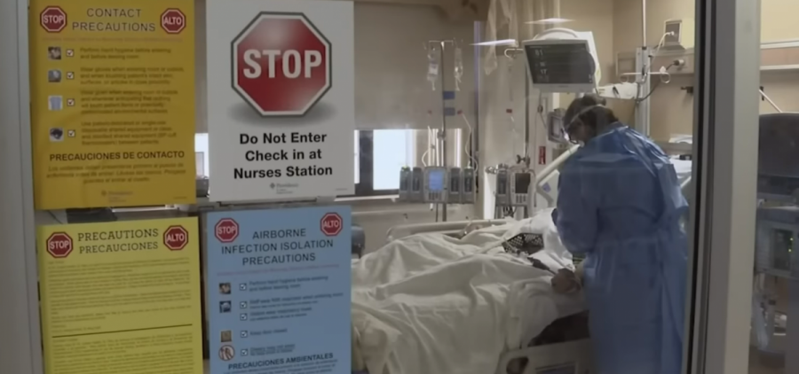 A nurse stands at the bed of a patient in a hospital. Signs on the window warn about precautions against infection disease.