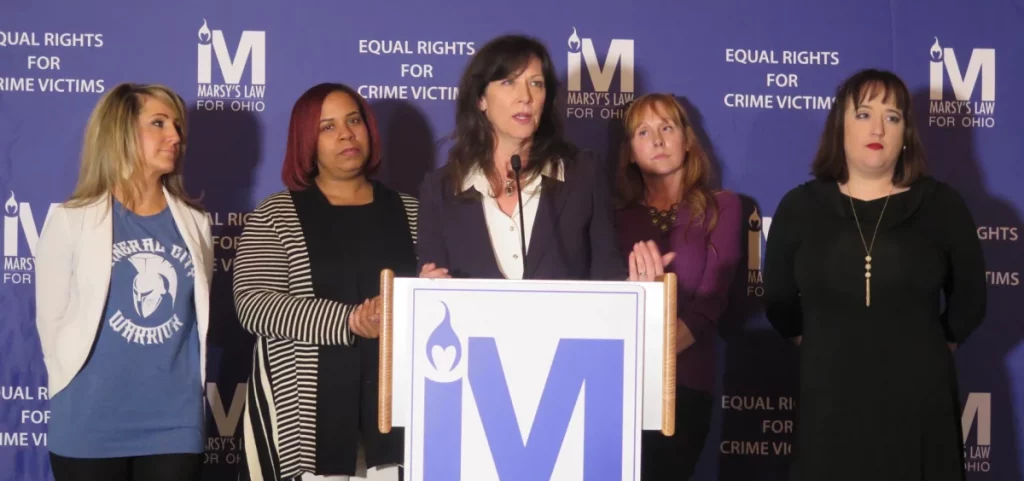 Supporters of Marsy's Law stand around a podium with the organization's logo on the front. They are standing in front of a backdrop with the logo and the words Equal Rights for Crime victims on it.