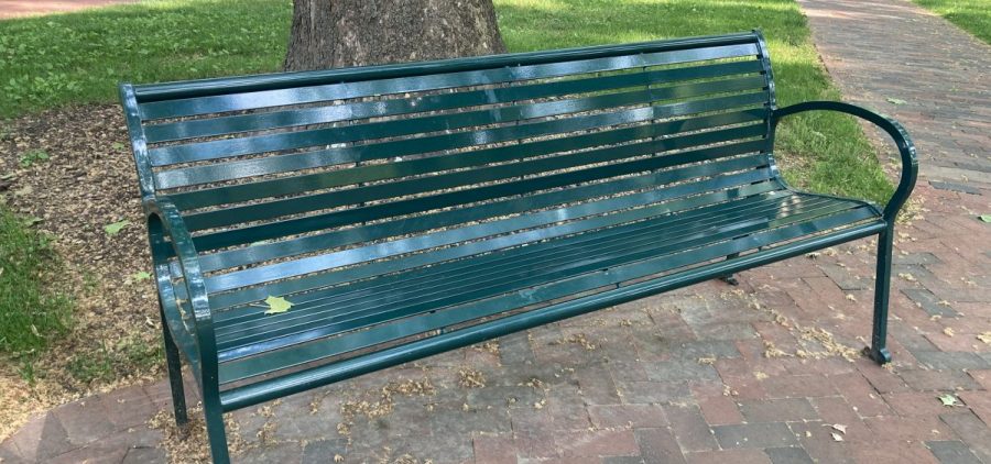A green metal bench sits on a brick path by a tree.