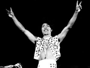 LITTLE RICHARD - US rock musician arms in air doing peace sign in glitter vest.