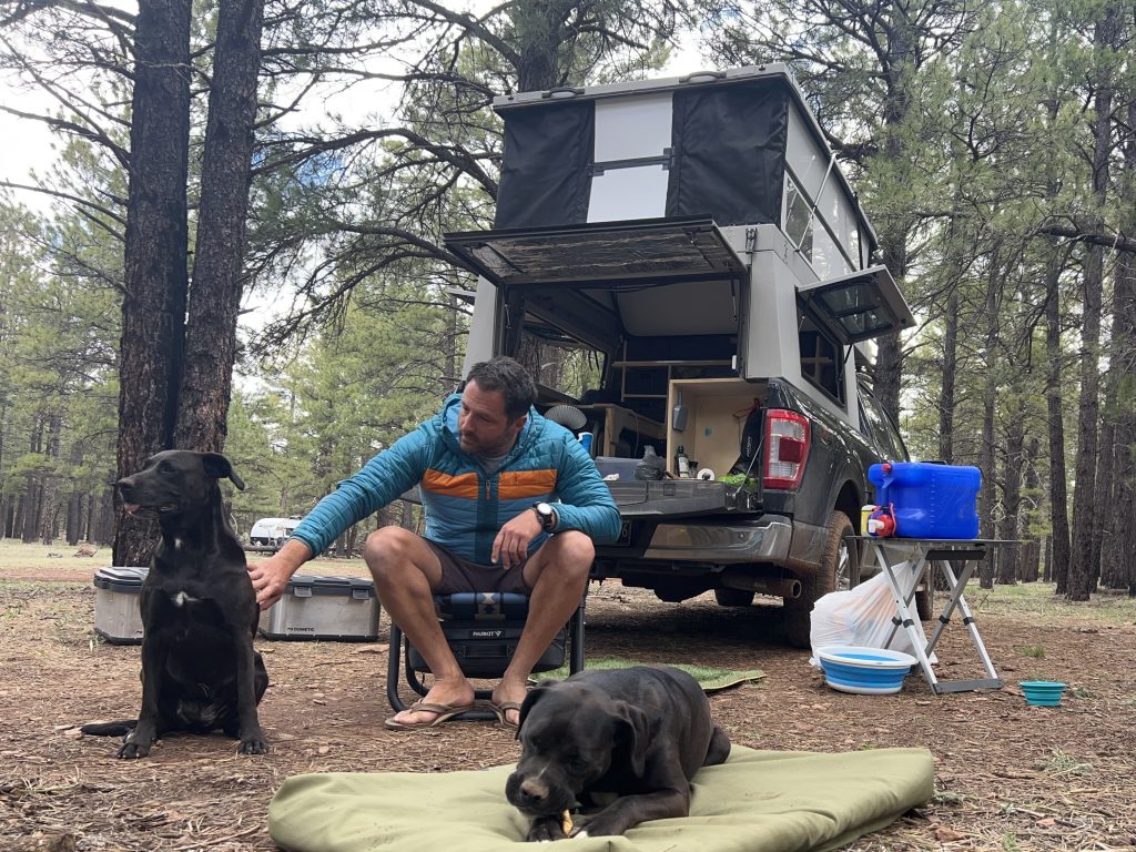 Veteran Jesse Reynolds sits in a camping chair petting one of his two dogs at a campsite in the woods.