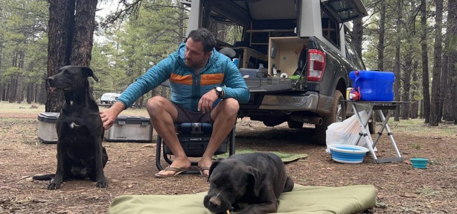 Veteran Jesse Reynolds sits in a camping chair petting one of his two dogs at a campsite in the woods.