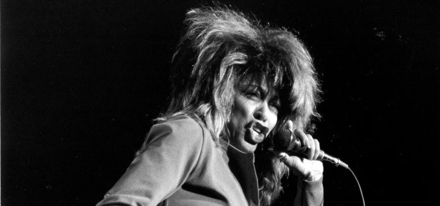 A black and white image of Tina Turner. She is wearing a short skirt and is singing into a microphone.