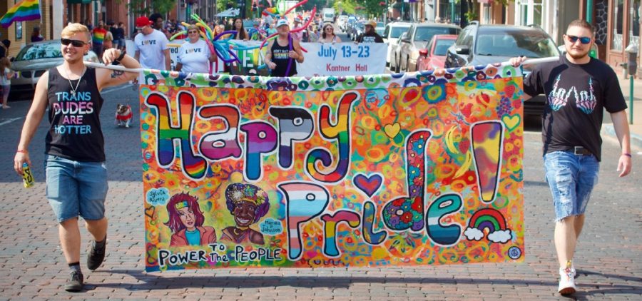 Two people holding a banner with the words “Happy Pride” in rainbow colors on a street with other people and buildings in the background.