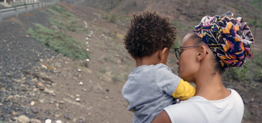 A Black woman tenderly cradles a child in her arms. Her colorful head wrap and sleek glasses frame her profile as she gazes down at the child, her expression filled with loving care. The child, clad in a light gray shirt over long yellow sleeves, rests peacefully on her shoulder, their face turned away from the viewer. In the backdrop is a rocky terrain with blurred railings and trees in the distance.
