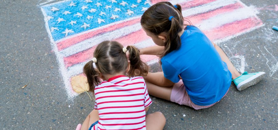 Two girls draw an American flag with colored chalks on the sidewalk near the house on sunny summer day.