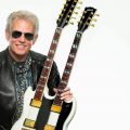 A promotional picture of guitarist Don Felder, holding a double neck guitar, wearing a scarf and a leather jacket - posed against a white background.