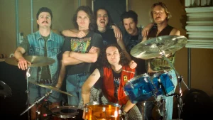 King Gizzard & the Lizard Wizard pose for a promotional shot. They are posed behind a drum set indoors. 