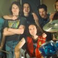 King Gizzard & the Lizard Wizard pose for a promotional shot. They are posed behind a drum set indoors.