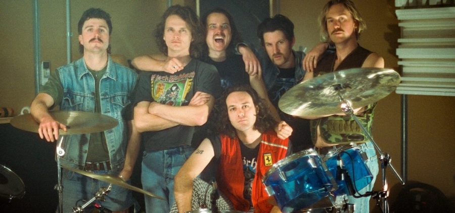 King Gizzard & the Lizard Wizard pose for a promotional shot. They are posed behind a drum set indoors.