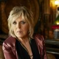 A promotional picture for musician Lucinda Williams. She is in a dark room and she has blonde shaggy hair and it wearing a red velvet blazer.