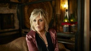 A promotional picture for musician Lucinda Williams. She is in a dark room and she has blonde shaggy hair and it wearing a red velvet blazer. 