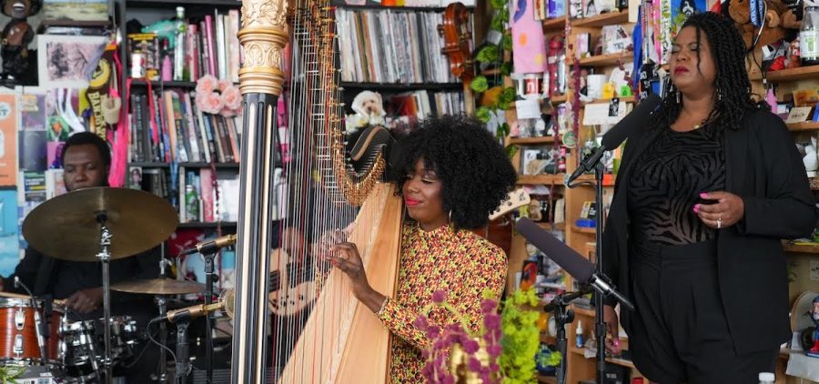 An image of Brandee Younger performing her Tiny Desk concert. She is playing a harp and wearing yellow, and she is flanked by a drummer on her left with a full kit and a singer on her right who is front of a microphone.