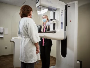 Two women, one in lab coat and one in a skirtsuit, stand in front of a diagnostic machine of some sort. 