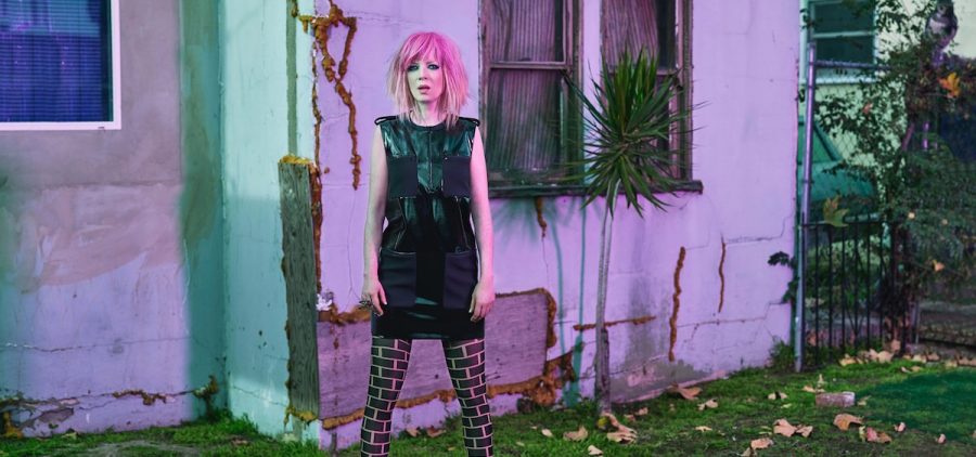 A promotional image for Garbage featuring the band's lead singer, Shirley Manson, with dyed pink hair and standing with her legs spread. She is wearing a black dress and there is a house with white siding behind her.