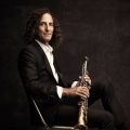 A promotional image of Kenny G. He is sitting in a chair with his saxophone against a black background.