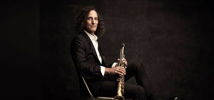 A promotional image of Kenny G. He is sitting in a chair with his saxophone against a black background.