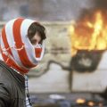 Northern Ireland youth stands against a background of a blazing van, his head covered in a striped scarf to protect his identity on May 7, 1981