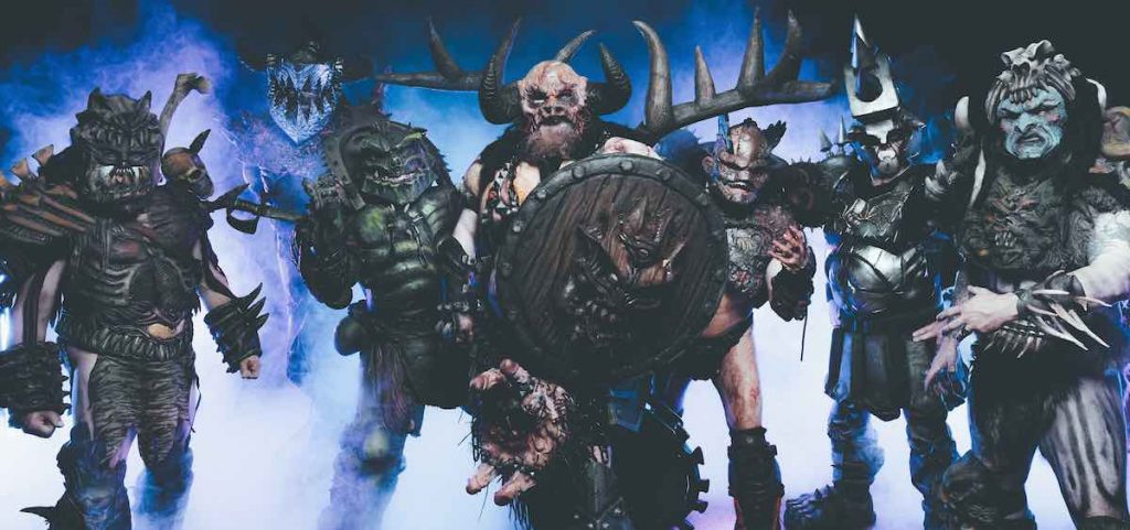 A promotional image for the band GWAR. The group wears over-the-top science fiction inspired costumes which include elaborate makeup and prosthetics. Six members of the band pose against a blue smoky background. 