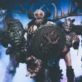 A promotional image for the band GWAR. The group wears over-the-top science fiction inspired costumes which include elaborate makeup and prosthetics. Six members of the band pose against a blue smoky background.