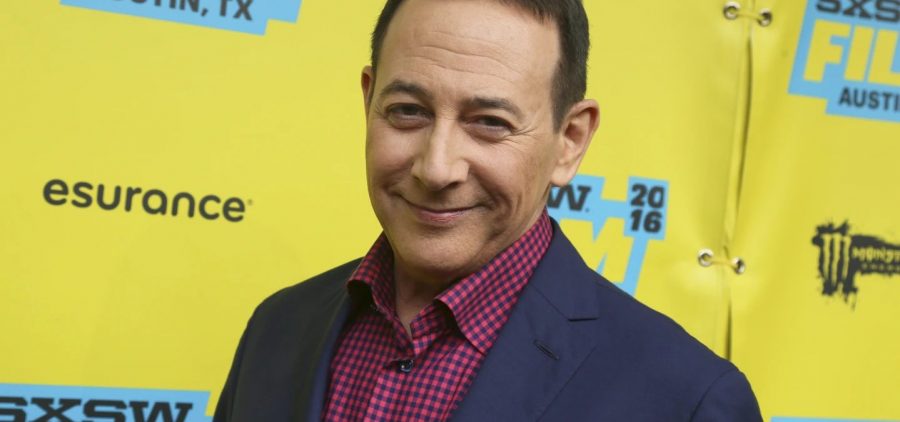 A picture of actor Paul Reubens. He is wearing a suit and is posed against a yellow background.