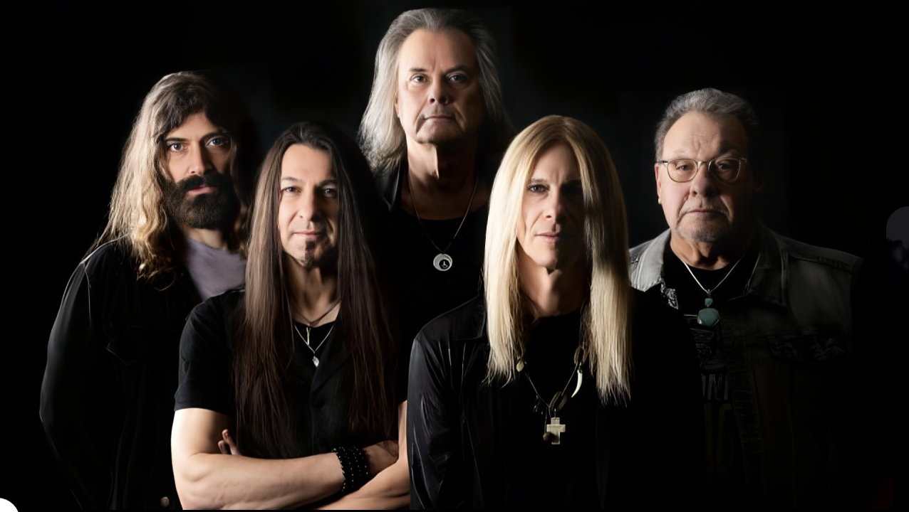 A promotional image of the band The Guess Who. There are five members in the band, and they are all wearing black and are posed against a black background. You can only see them from the torso up.