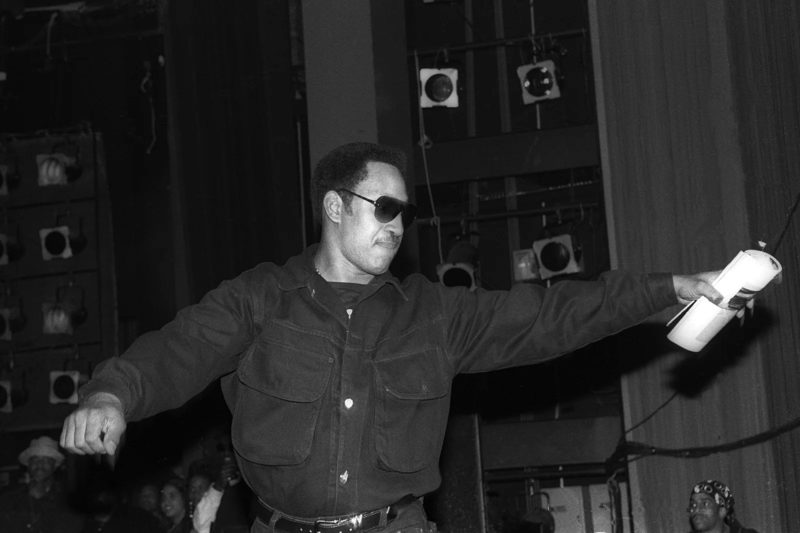 A black and white image of DJ Kool Herc. He is wearing sunglasses and behind him there is a stand of speakers. 