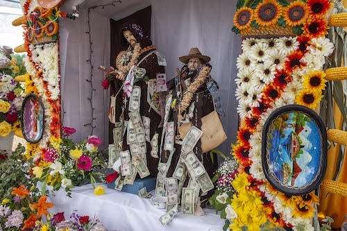 Two bearded statuettes, wearing long brown gowns and decorated with dollars, pesos and cookie necklaces, sit on a white altar covered in colorful flowers, artwork and corn.