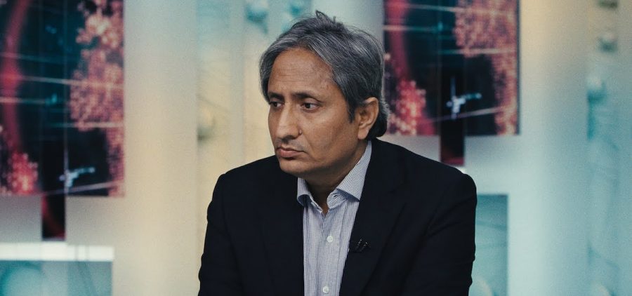 Ravish Kumar stands in the center of the image, dressed in a dark blazer and patterned dress shirt, looking off with a worried expression. The blurred background creates an abstract effect, making it difficult to discern the shapes and colors behind him. Despite his sharp attire, his demeanor and posture suggest a sense of concern.