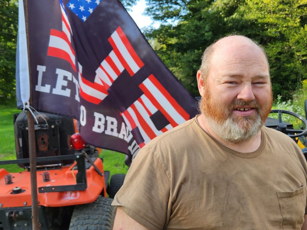 James Batchelder of West Topsham, Vermont poses for a portrait in front of a tractor with a let's go brandon flag on it.