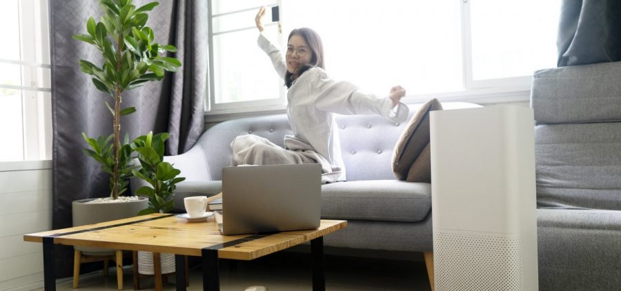 A woman in a robe stretches with a smile while sitting on a couch. There is an air purifier in the room near her.