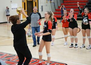 Alexander Spartan Lexi Grissett smiles with her head coach who holds up the golden volleyball for Grissett's milestone. Spartan teammates cheer in the background.