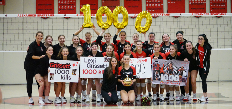 Alexander high school volleyball senior Lexi Grissett sits with her team in a group photo with numbered balloons and a golden ball to celebrate her 1,000th kill achievement