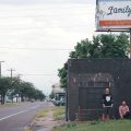 A promotional image of the band Chat Pile. They are pictured from far away, under a billboard sign that is chipped and aged.