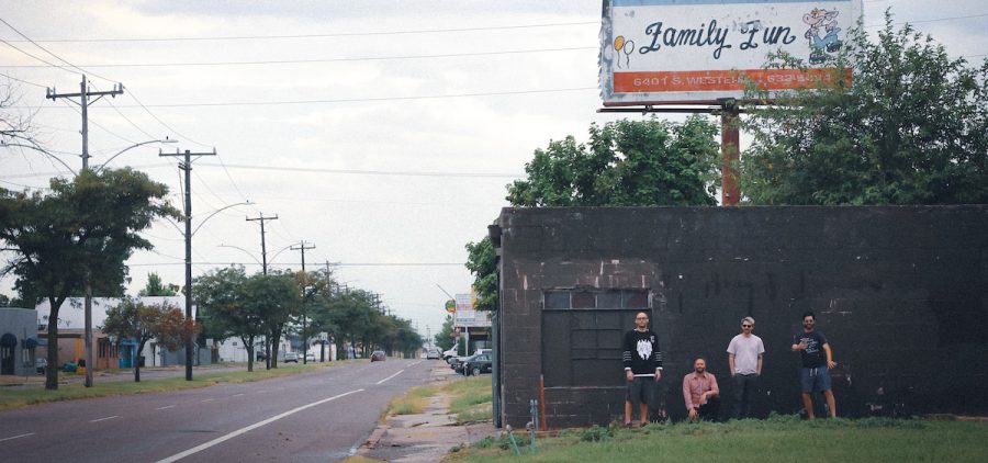A promotional image of the band Chat Pile. They are pictured from far away, under a billboard sign that is chipped and aged.