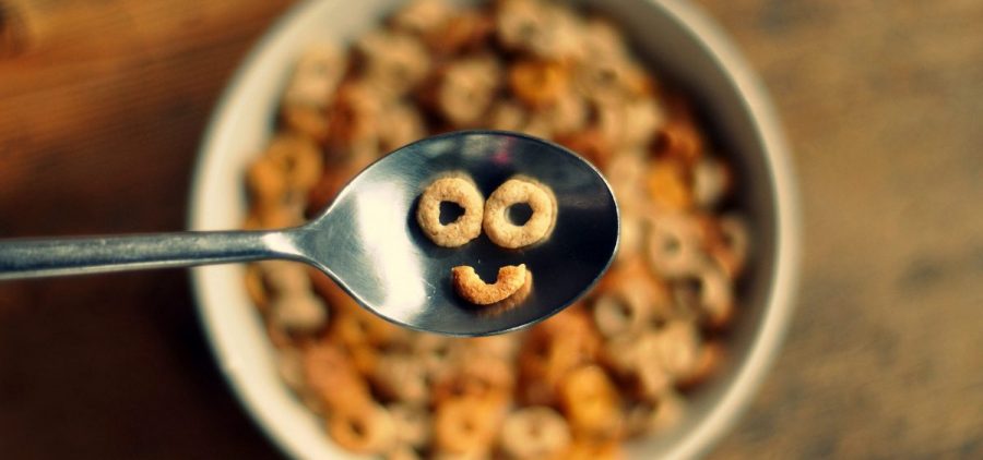 A spoon full of circular cereal has three pieces that form a smiley face over a bowl of the same cereal.