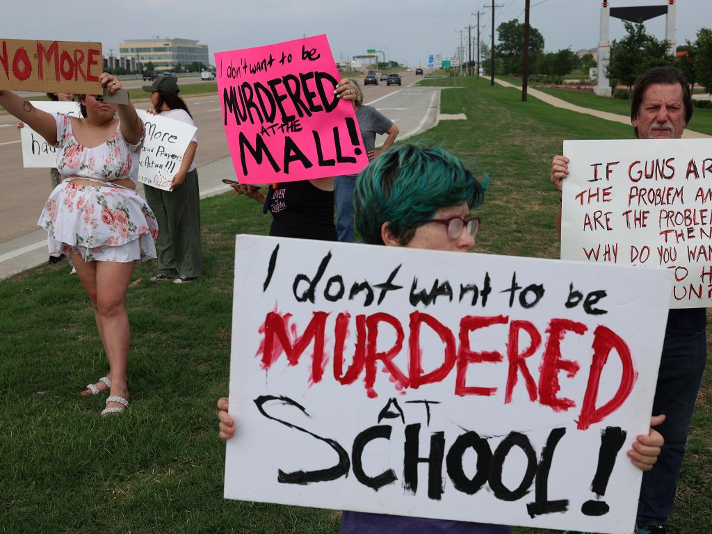 Children hold up signs protesting gun violence along a road. The child in front has a white sign with black and red lettering that reads I don't want to be murdered at school.