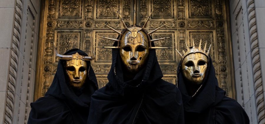 A press photo for the heavy metal band Imperial Triumphant. The three band memebrs are wearing gold masks that are in an art deco style with black robes.