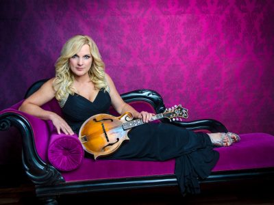 A press image of bluegrass musician Rhonda Vincent. She is wearing a black dress and leaning on a purple couch.