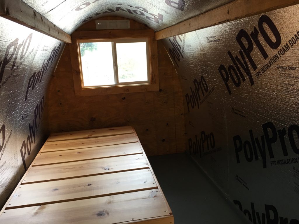 The inside of the Conestoga hut. There is a wooden bed frame that takes up half the floor and a window. The insulation reinforcing the tarp is visible.