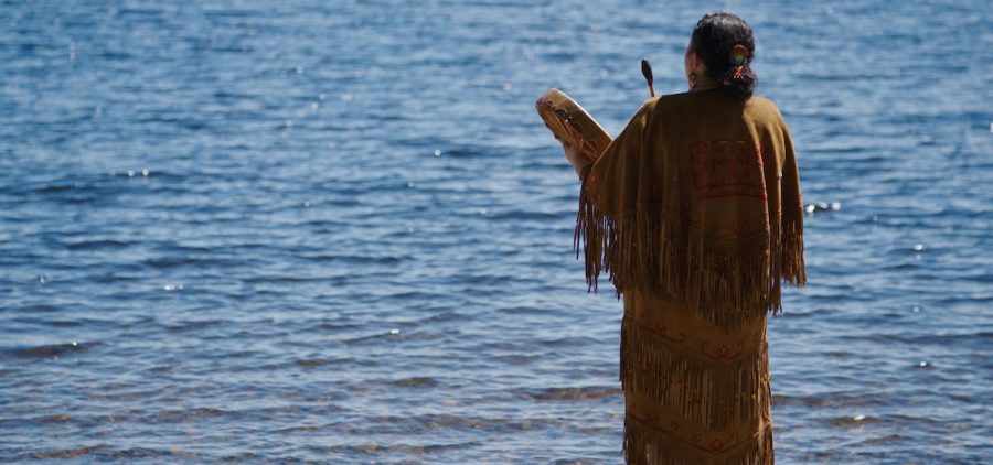 Lauren Stevens, a member of the Passamaquoddy tribe, back to the camera, playing a drum and singing on the shore of Pine Island.