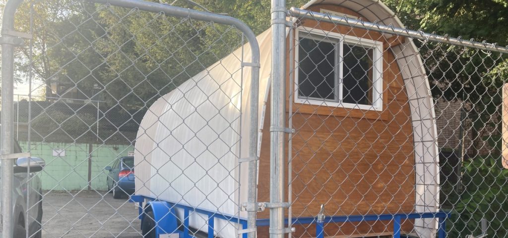 A Conestoga hut with a wood back, a small elevated window, and a tarp reinforced with wiring and insulation for covering.