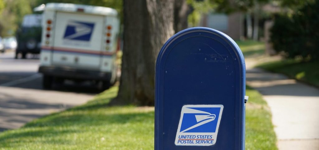 A United States Postal Service mailbox stands along a sidewalk with a mail truck off in the distance.