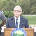 Ohio Governor Mike DeWine talks about new distracted driving law in Delaware county at a podium with a state trooper over one shoulder and some older looking white guy in a suit over his right shoulder.