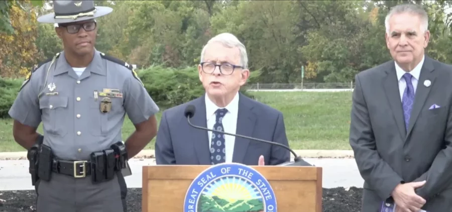 Ohio Governor Mike DeWine talks about new distracted driving law in Delaware county at a podium with a state trooper over one shoulder and some older looking white guy in a suit over his right shoulder.