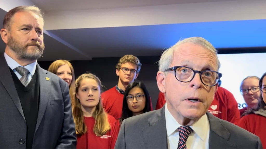 Ohio Governor Mike DeWine speaks to reporters as Attorney General Dave Yost and student volunteers look on.