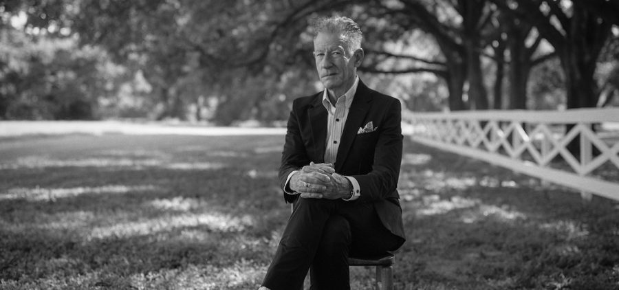 A promotional picture of Lyle Lovett. It is black and white and Lyle is sitting underneath trees that cast shadows over him.